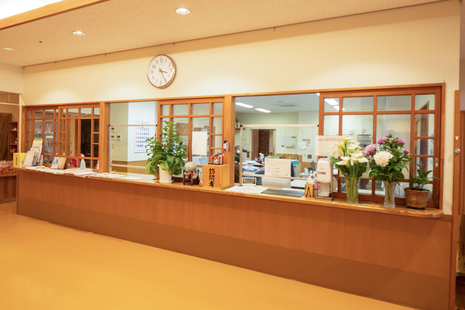 受付カウンター。木製フレームのガラス張り受付、観葉植物、壁時計のある明るい施設エントランス