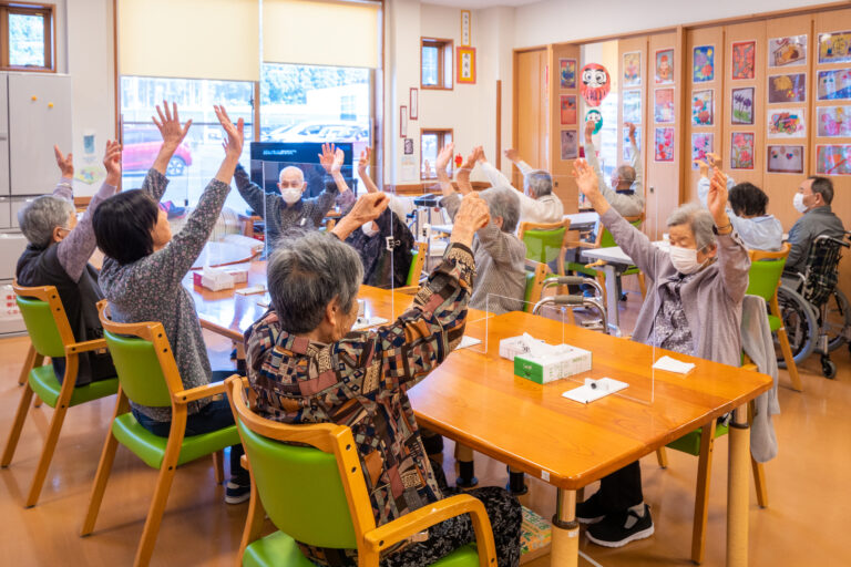 介護施設でのレクリエーション。高齢者たちが手を上げて楽しく参加している様子。